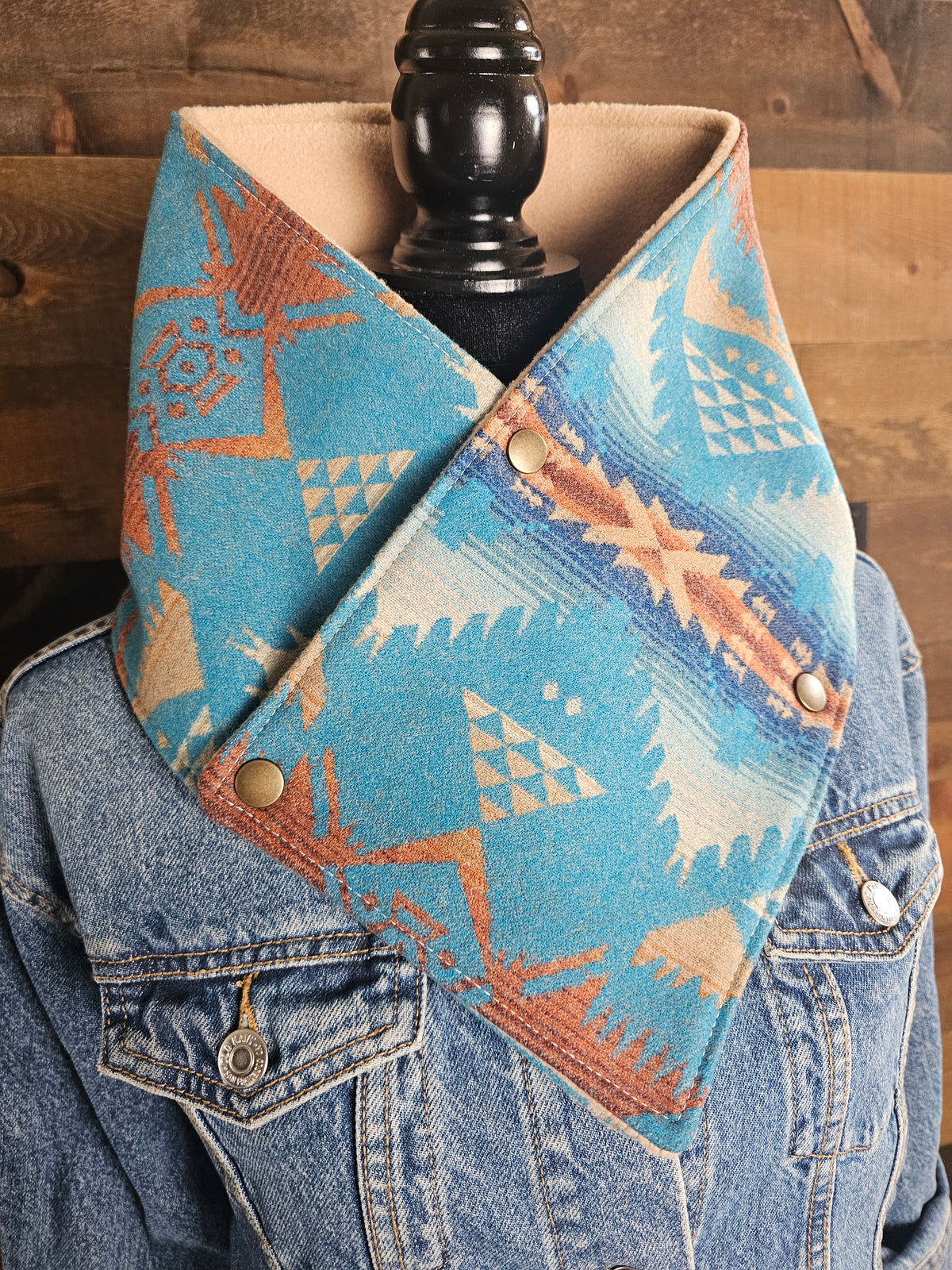 Close-up of the turquoise Nehalem cowl on denim, showing cream geometric motifs, rust accents, and snap closures.