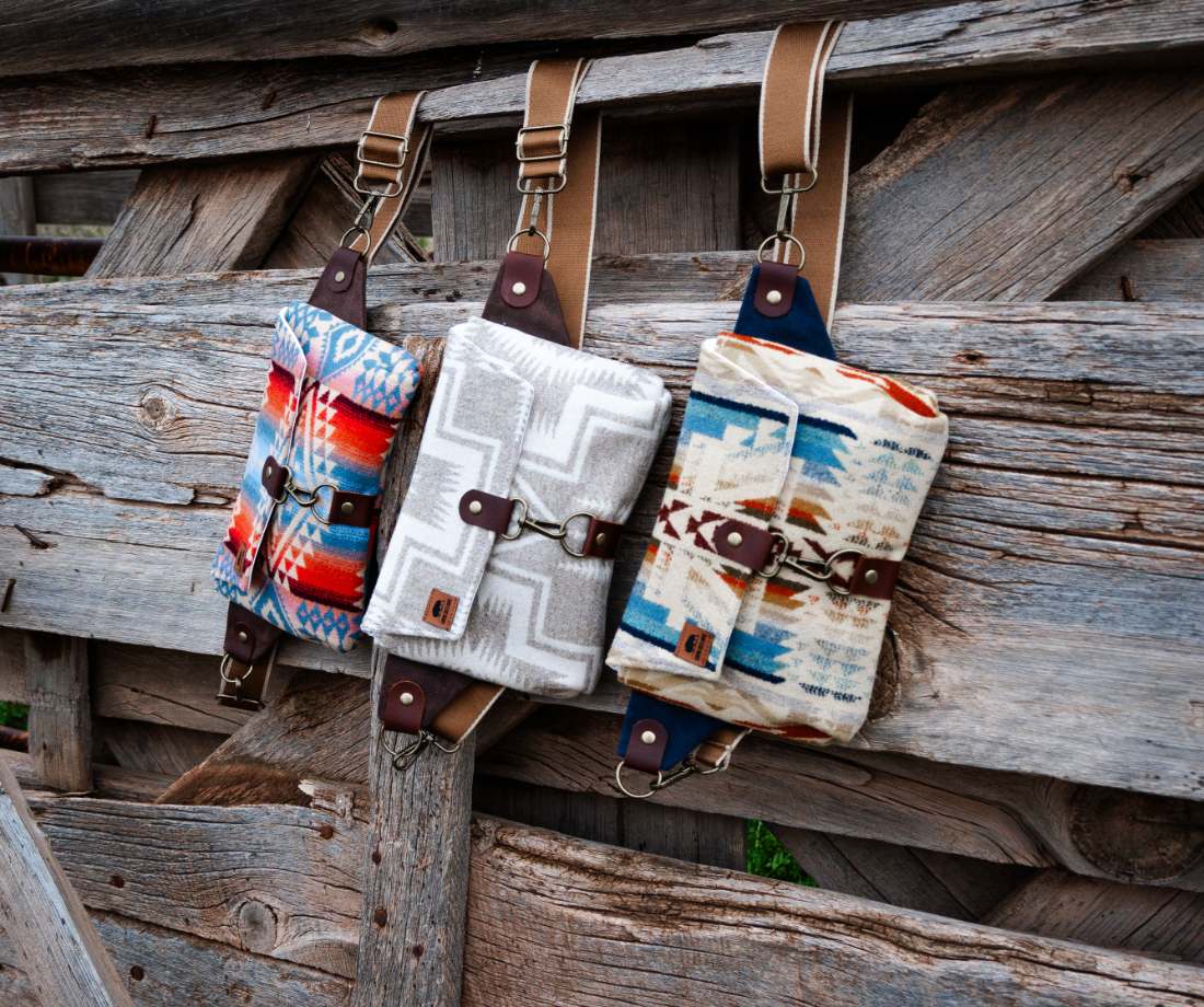 Three patterned wool sling bags with tan straps hanging on a weathered wooden fence.
