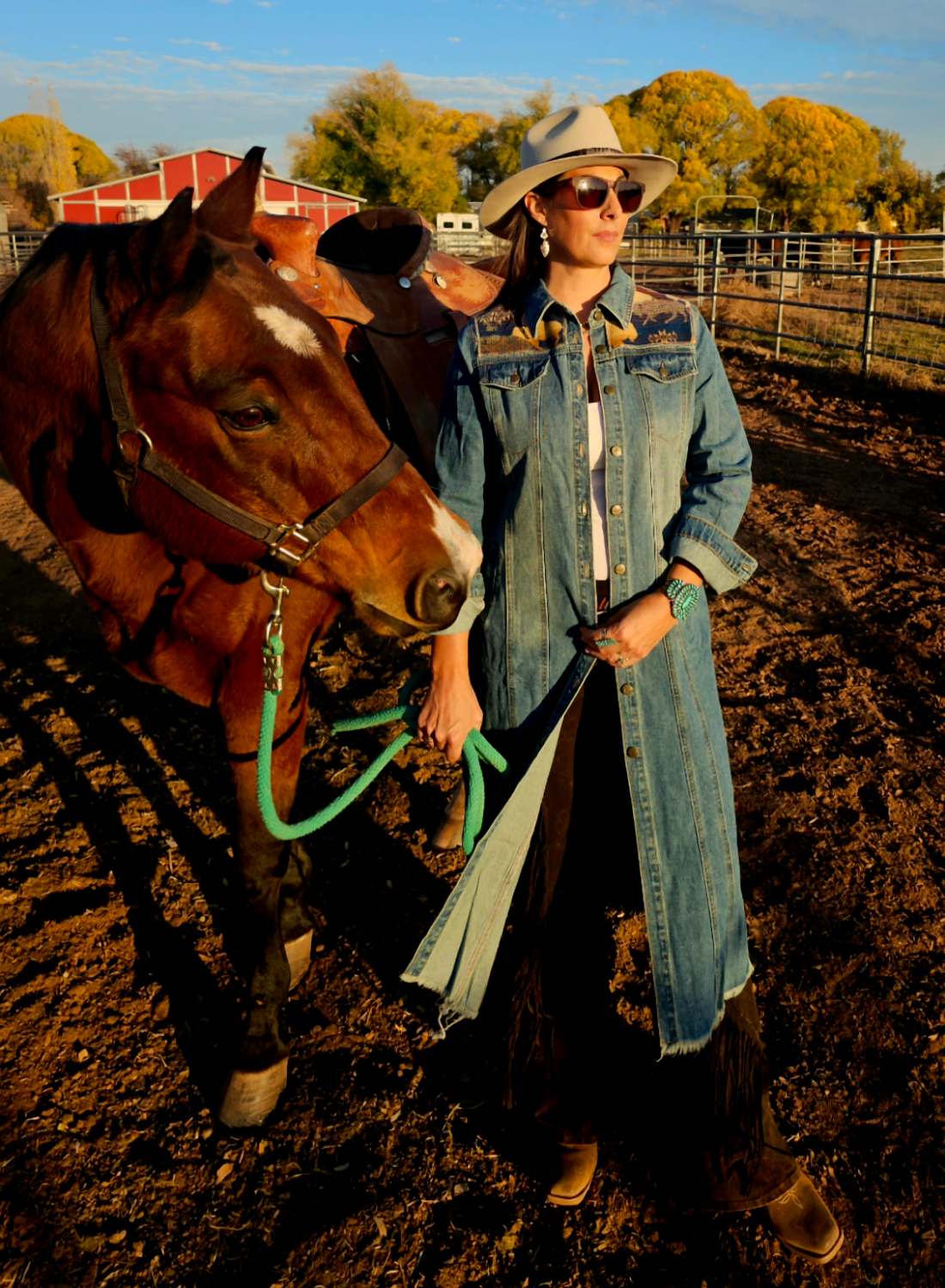 Woman wearing a long denim coat and wide-brimmed hat beside a saddled brown horse.