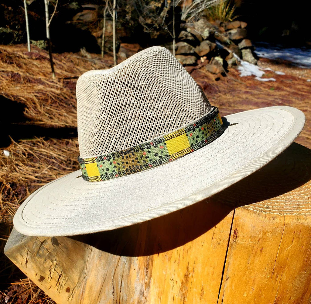 Monster Brown trout-patterned hat band wrapped around a light mesh-crown hat resting on a tree stump.
