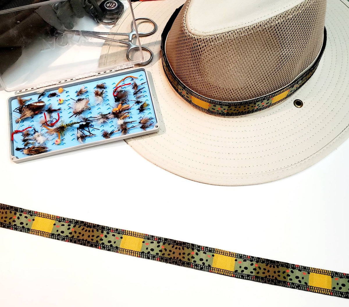 Monster Brown trout hat band laid straight beside a light hat and fishing fly box.