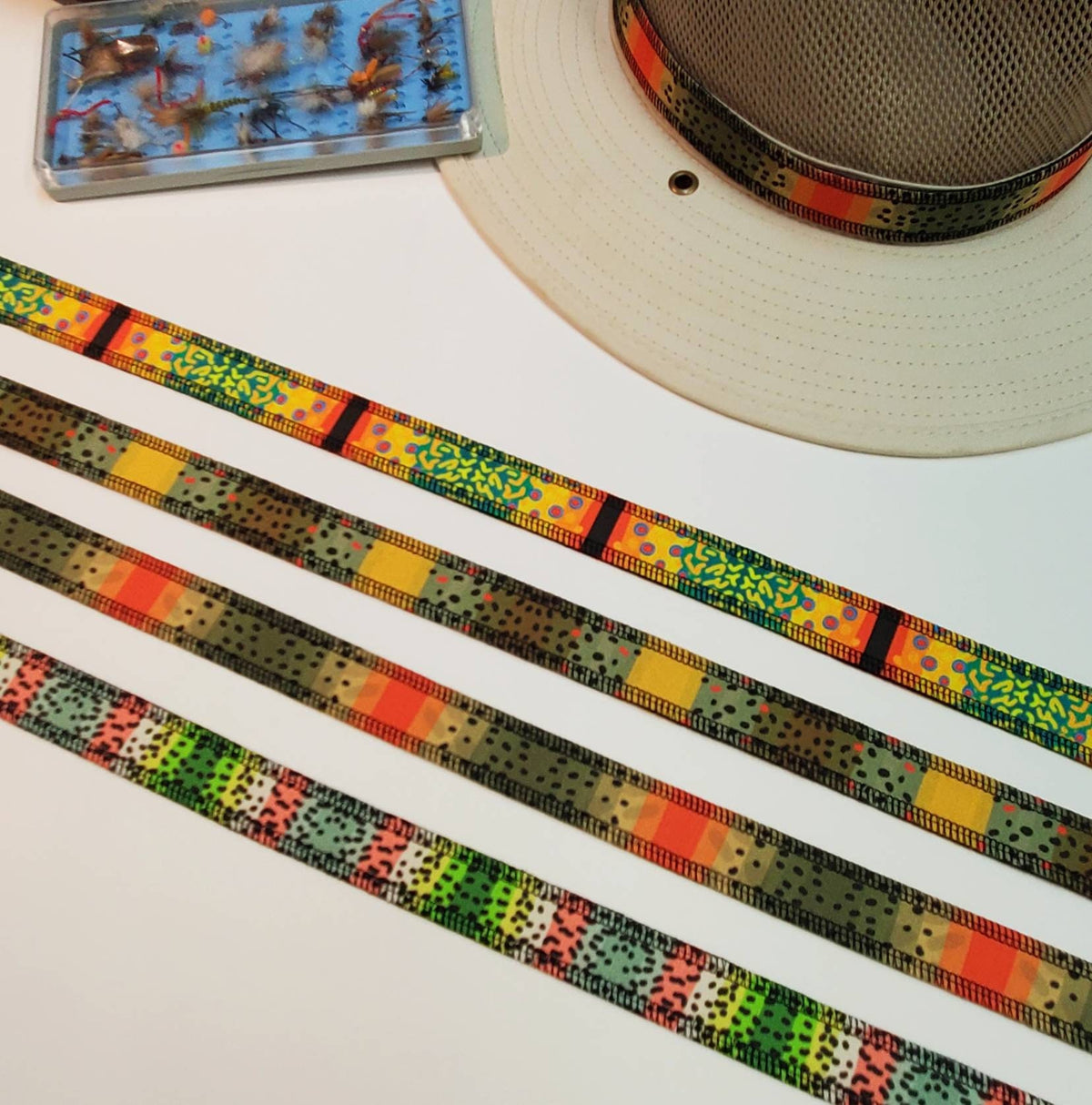 Four trout-patterned hat bands laid side by side beside a hat and fishing fly box.