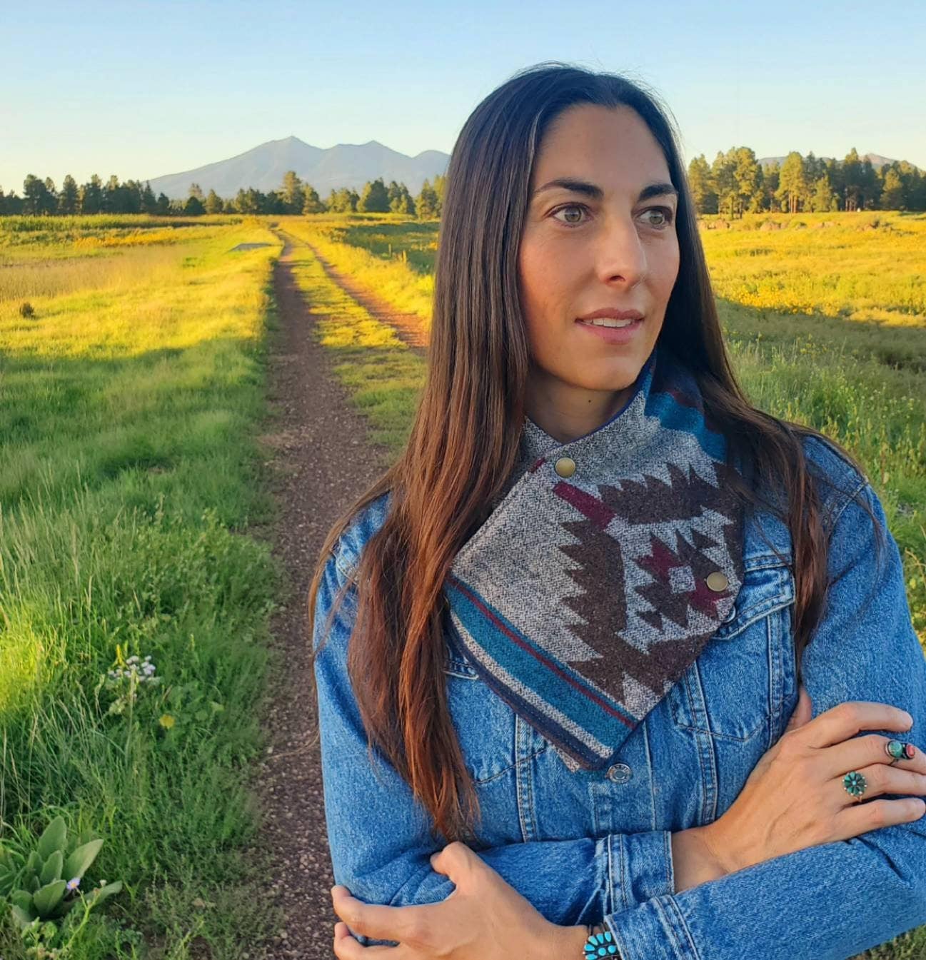 Woman standing in grass field, modelling a wool cowl scarf wrapped around her neck. Scarf features southwestern motif with blue, brown and magenta hues and three antique brass snaps.