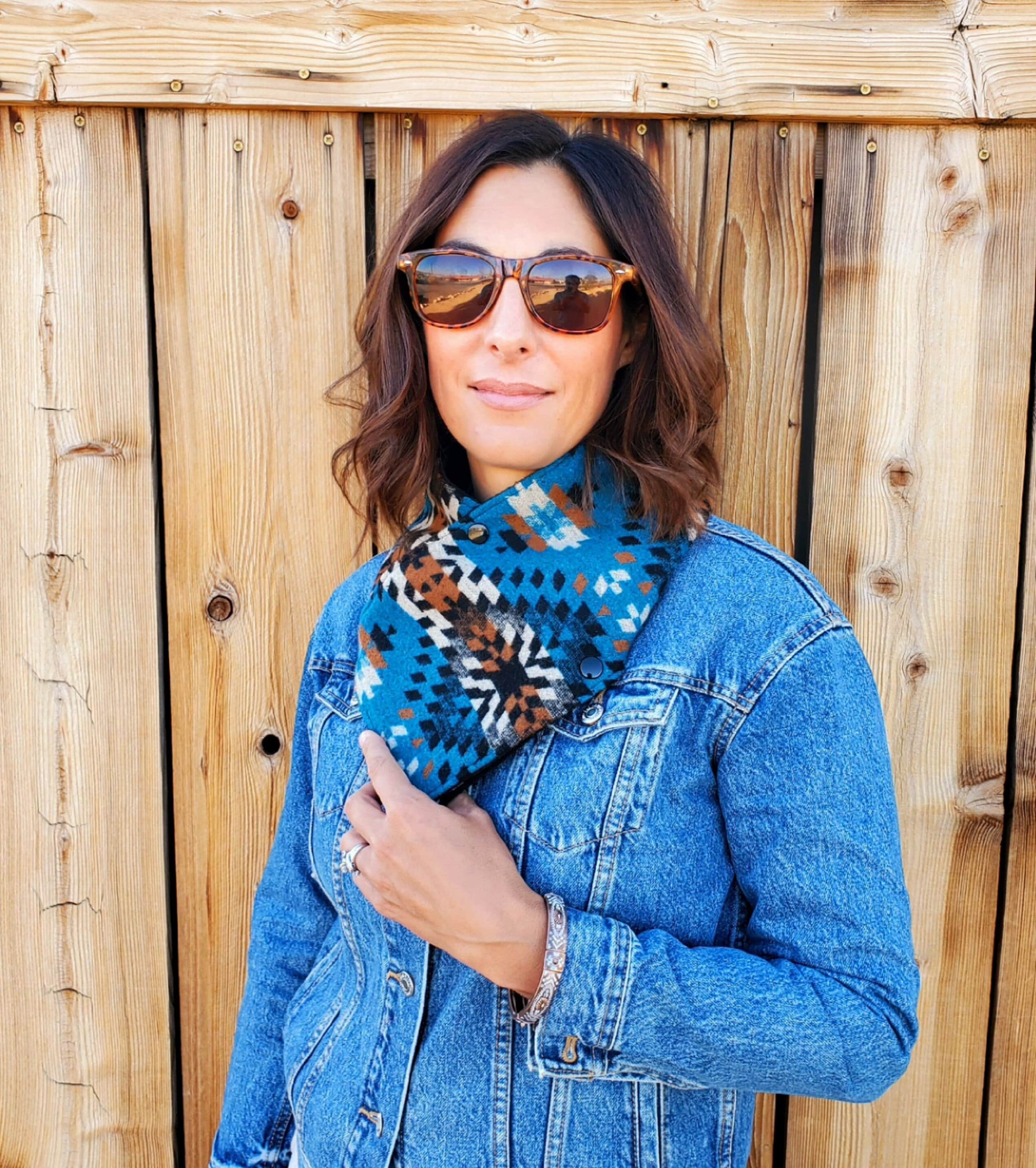 Model is standing in front of wooden door, wearing western cowl scarf. Bandana scarf is made from wool & cotton, features blue, white, black & brown geometric designs.