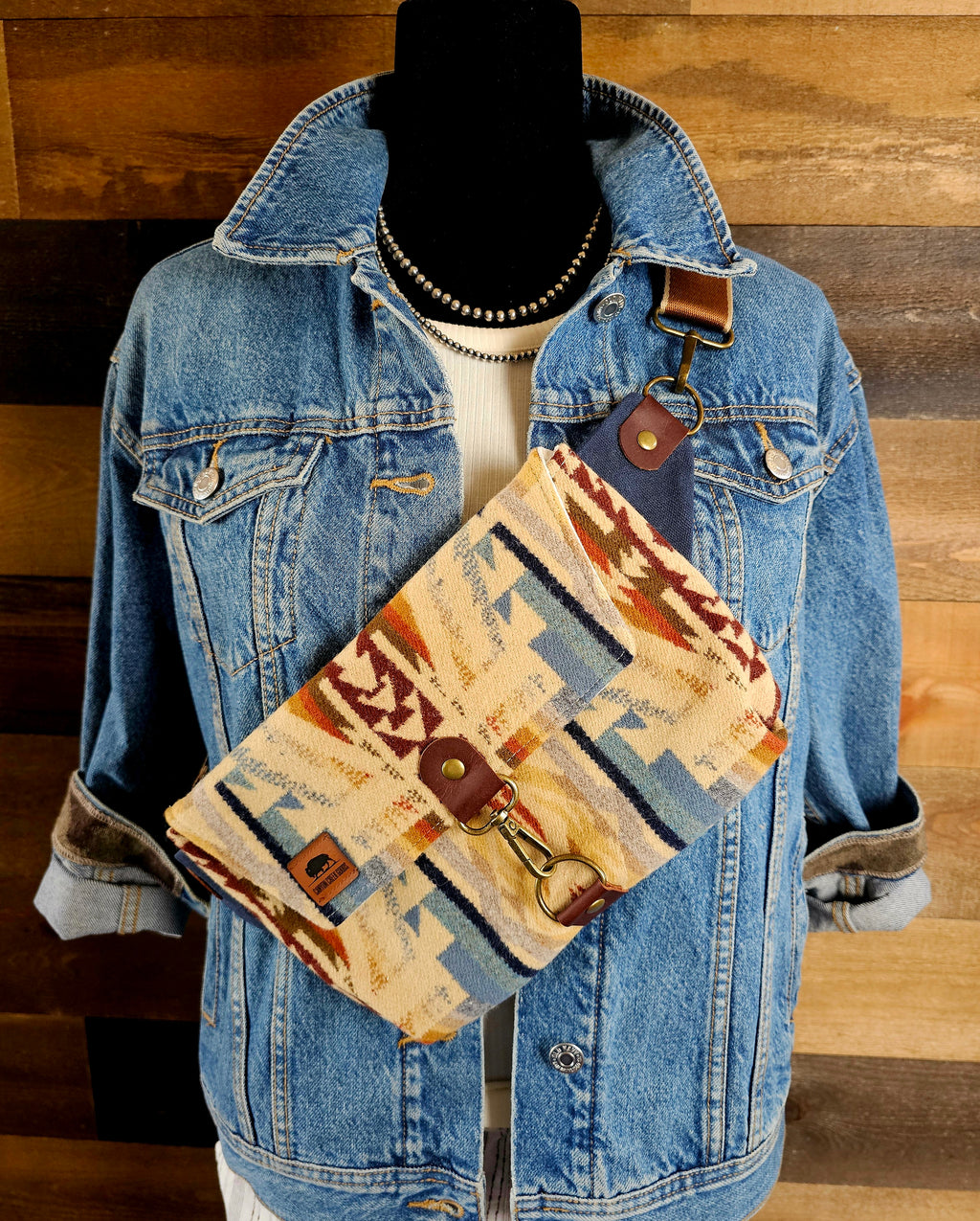 Shonto Wheatlands crossbody sling worn over a denim jacket, with tan, blue and rust patterned fabric and a brown strap.