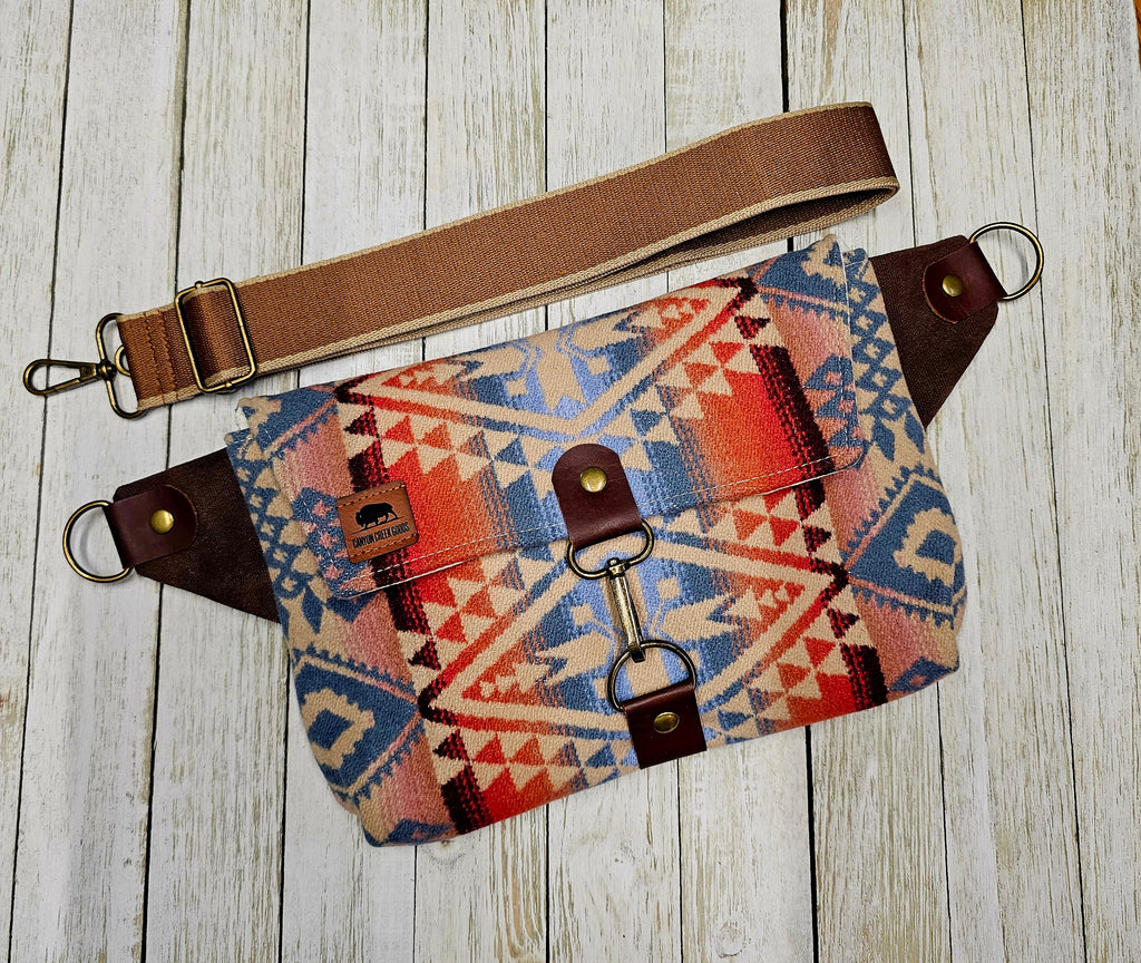 Canyonlands sling laid flat, showing its colorful geometric pattern, exterior zipper and brown strap.